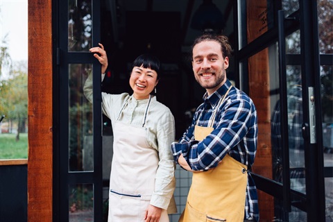 Male and female wearing aprons and smiling while standing at the front of their cafe