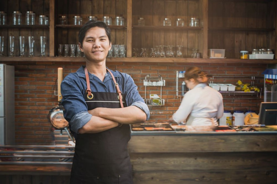 Main in apron posing in front of restaurant kitchen.