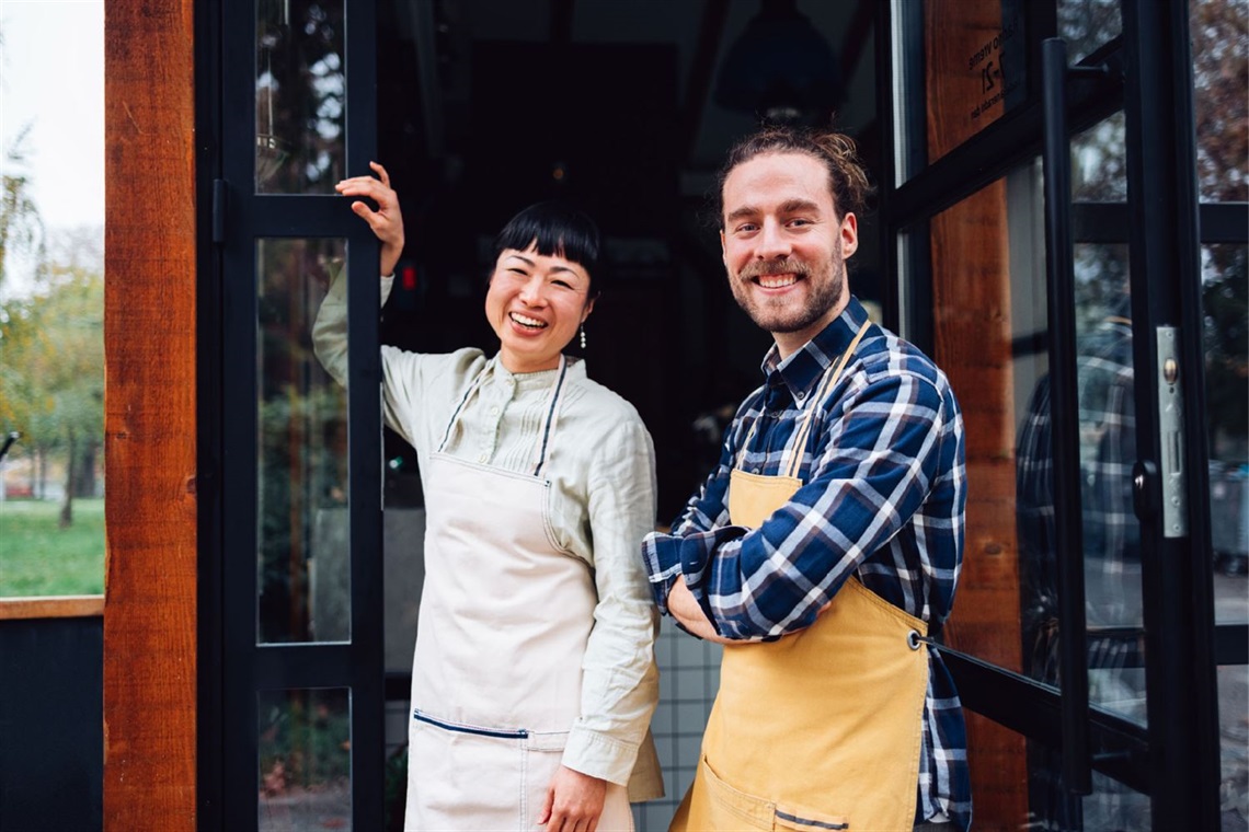 Male and female wearing aprons and smiling while standing at the front of their cafe.