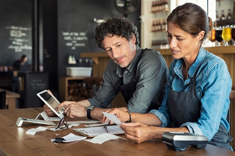 Male and female wearing aprons sitting at a table working out finances with a calculator, EFTPOS machine and tablet