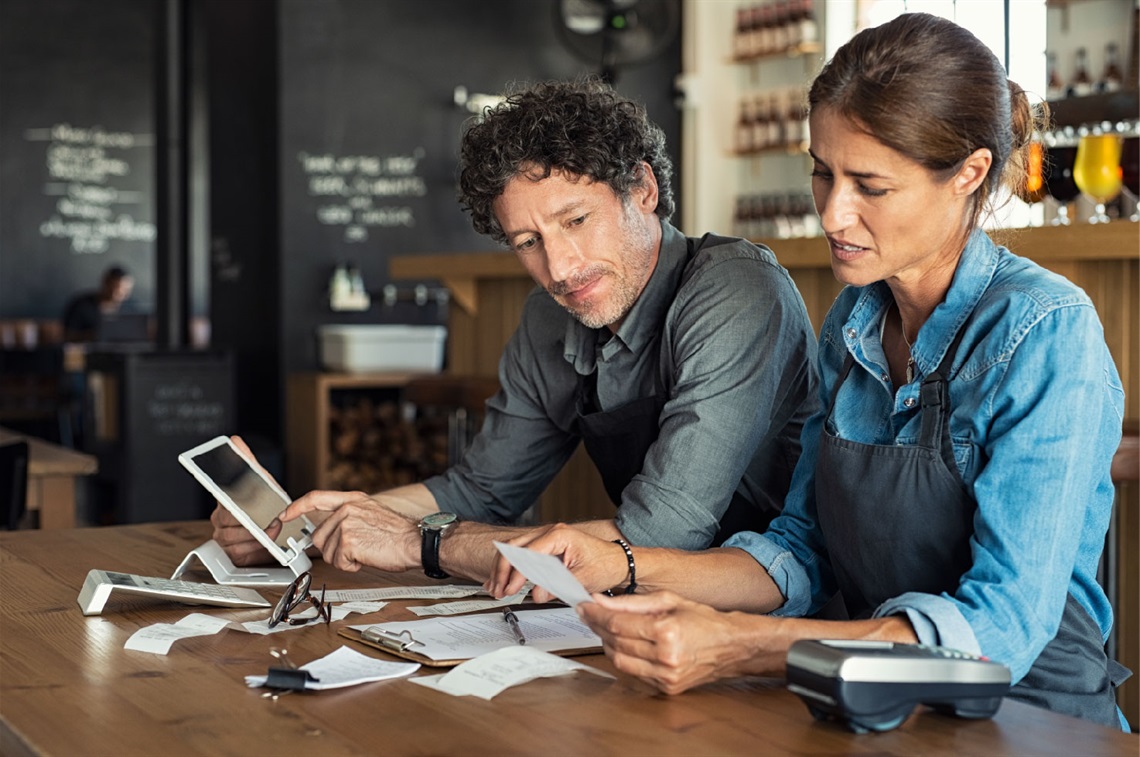 Male and female wearing aprons sitting at a table working out finances with a calculator, EFTPOS machine and tablet.