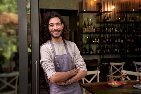 Man in apron standing at the entrance to his restaurant with tables, chairs and shelves visible in the background