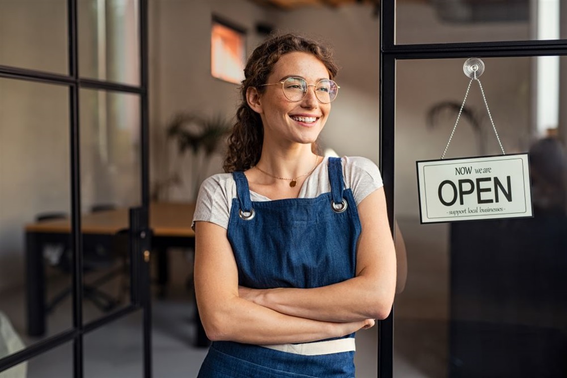 Young lady in glasses standing next to a sign which says 'now we are open - support local businesses'.